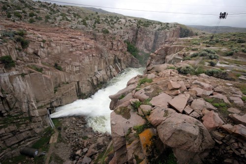 Aerial view of a river flowing through a rocky canyon, with a dam releasing water and a cable car overhead.