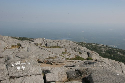 A rocky mountain summit with a view of distant hills and a sign pointing to "State Park HQ."