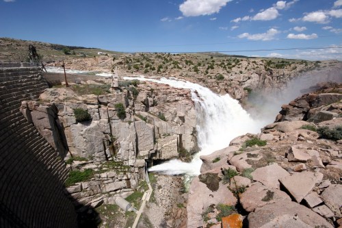 A powerful waterfall cascades over rocky cliffs, with a dam visible on the left and a blue sky above.