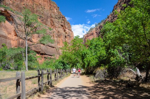 Two people walk along a dirt path surrounded by tall red rock formations and lush green trees under a blue sky.