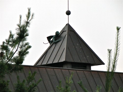 A bronze monkey sculpture perched on the peak of a sloped roof, surrounded by pine trees.