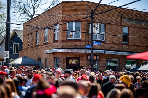 A large crowd gathers outside a brick building on a sunny day, with signs and tents visible in the background.