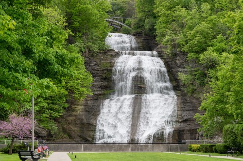 A cascading waterfall surrounded by lush green trees, with a park bench in the foreground and a footbridge above.
