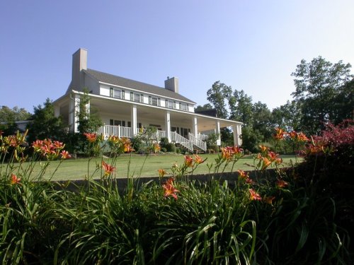 A large white house with a porch, surrounded by vibrant flowers and greenery under a clear blue sky.