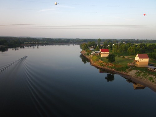 A serene river scene with a boat, green trees, and houses reflecting on the water under a clear sky.
