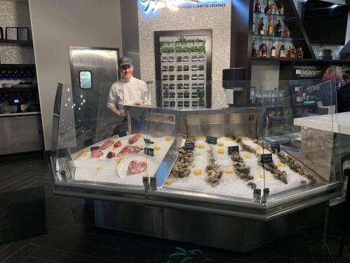 A seafood display featuring oysters and various cuts of meat, with a smiling chef behind the counter.