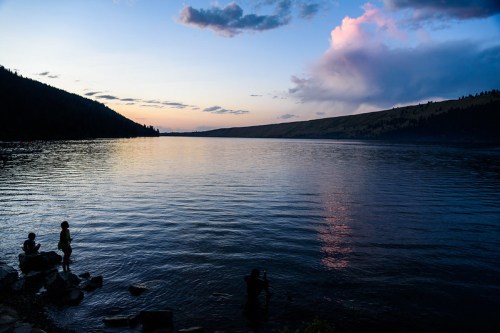 Silhouettes of people by a calm lake at sunset, with colorful clouds reflecting on the water's surface.