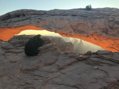 A natural rock arch frames a scenic view of distant cliffs and a sunset, with sparse vegetation on the arch's surface.
