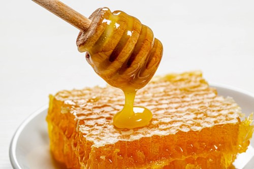 A honey dipper drizzles golden honey over a piece of honeycomb on a white plate.