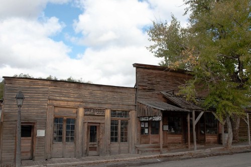 A rustic wooden building in a ghost town, featuring signs for "Dry Goods" and "Tobacco & Cigars," under a cloudy sky.