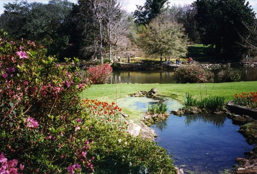 A serene landscape featuring a pond, colorful flowers, and trees, with people walking along a path in the background.