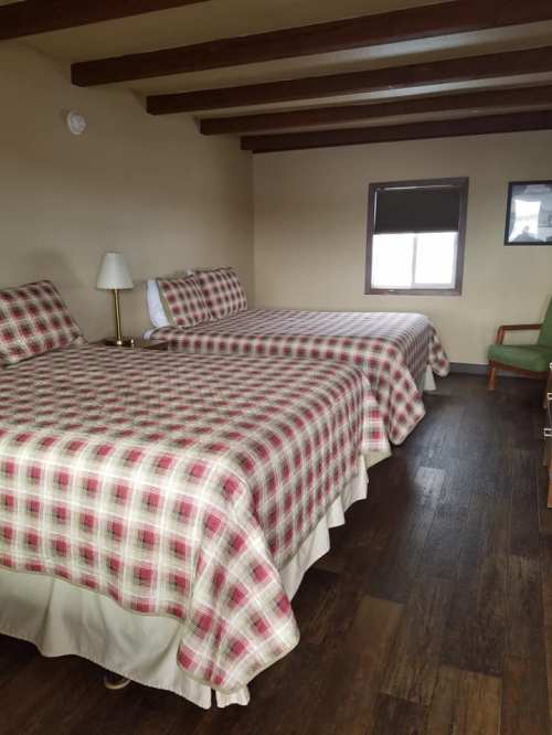 A cozy room with two plaid-covered beds, a lamp, a window, and a green chair. Wooden beams on the ceiling.