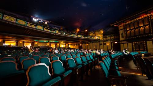 A spacious theater with teal seats, dim lighting, and a few audience members scattered throughout.