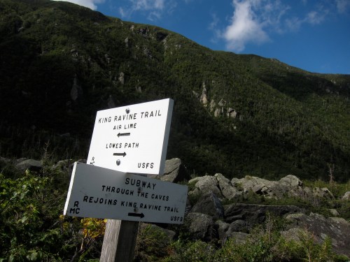 A trail sign for King Ravine Trail and Subway, surrounded by lush green mountains and a clear blue sky.