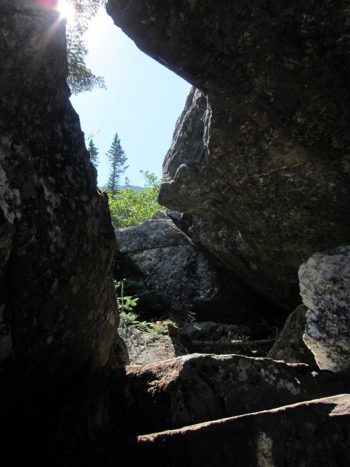 A view through a narrow gap between large rocks, with trees and a bright sky visible in the background.