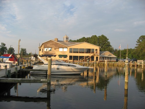 A waterfront building with a wooden deck, surrounded by boats and trees, reflecting in calm water under a cloudy sky.