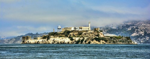 A distant view of Alcatraz Island, featuring its historic buildings and lighthouse against a cloudy sky.