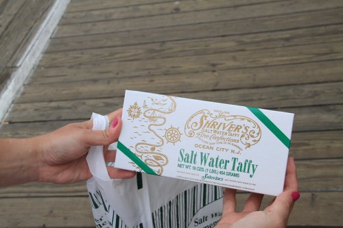 A person holds a box of Shriver's Salt Water Taffy, with a white and green design, on a wooden surface.