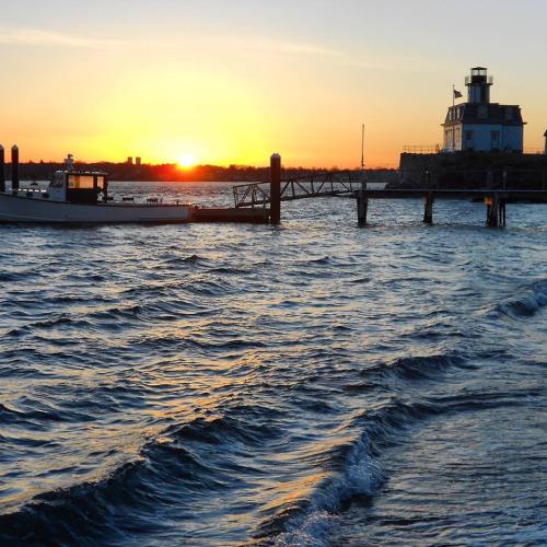 A serene sunset over water, with a boat near a pier and a lighthouse in the background. Waves gently ripple in the foreground.