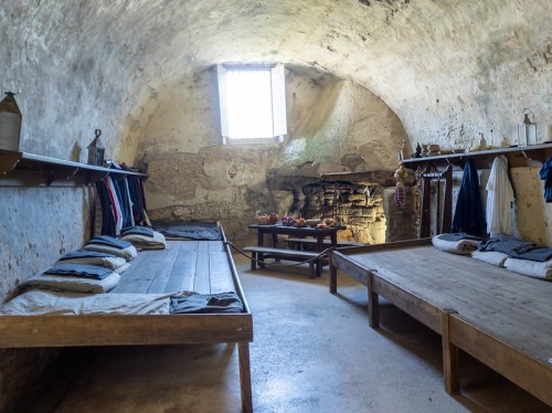 A rustic room with two wooden beds, shelves with clothes, and a table with food, set in a stone-walled space.