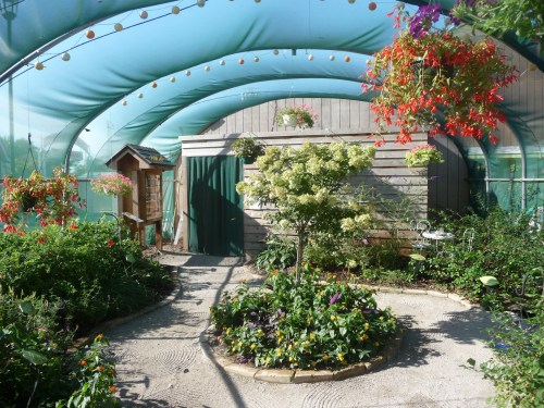 A vibrant greenhouse with hanging plants, a small shed, and a circular garden path surrounded by colorful flowers.