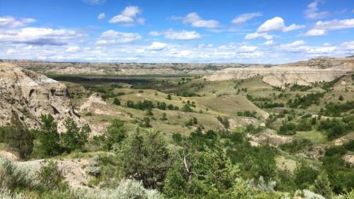 A panoramic view of rolling hills and valleys under a blue sky with fluffy clouds, dotted with green vegetation.
