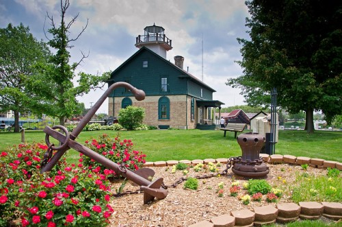 A historic lighthouse with a green exterior, surrounded by flowers and an anchor in the foreground.