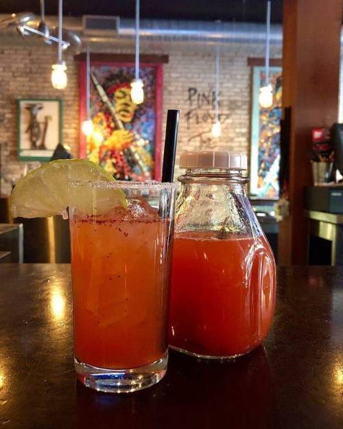 A vibrant cocktail with a lime wedge next to a small glass bottle of juice, set in a cozy bar with artistic decor.