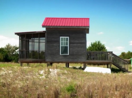 A small wooden house with a red roof, featuring a porch and surrounded by grassy land under a clear blue sky.