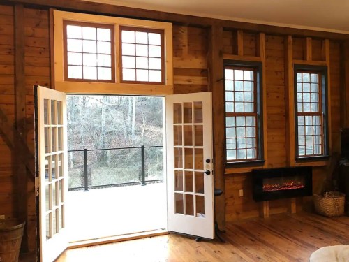 A cozy wooden room with large windows and double doors opening to a balcony, featuring a modern electric fireplace.