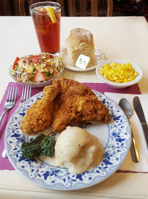 A plate of fried chicken with mashed potatoes and gravy, corn, a salad, a biscuit, and a drink on a table.