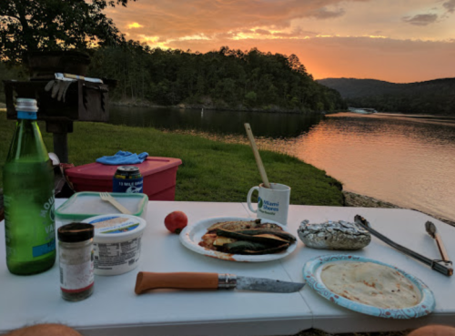 A table set by a lake at sunset, featuring food, drinks, and cooking utensils with a scenic view in the background.