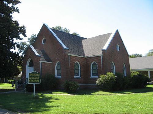 A brick church with arched windows, surrounded by green grass and bushes, under a clear blue sky.