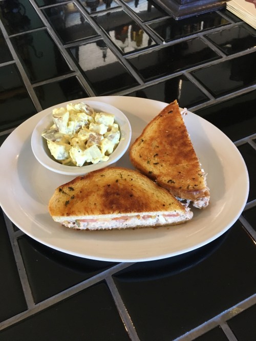 A toasted sandwich cut in half, served with a side of potato salad in a small bowl on a white plate.
