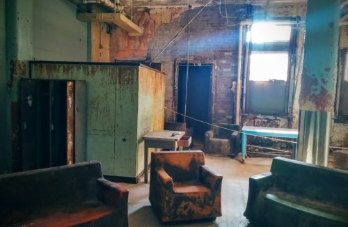 Abandoned room with rusty furniture, lockers, and peeling walls, illuminated by natural light from windows.