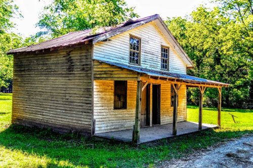 A rustic wooden house with a porch, surrounded by greenery and trees under a clear blue sky.