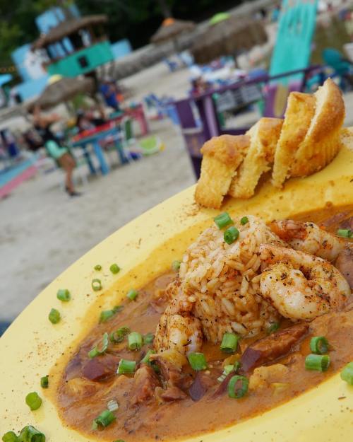 A vibrant plate of shrimp and rice garnished with green onions, served with cornbread, set against a beach backdrop.