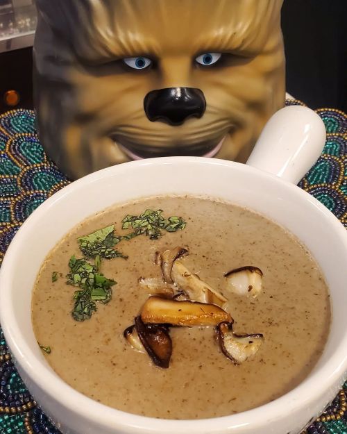 A bowl of creamy mushroom soup garnished with herbs, with a whimsical mask in the background.