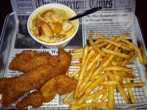 A tray with fried chicken tenders, French fries, and a cup of creamy soup on a newspaper background.