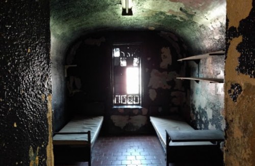 A dimly lit, empty prison cell with two beds and a barred window, featuring peeling walls and a stark atmosphere.