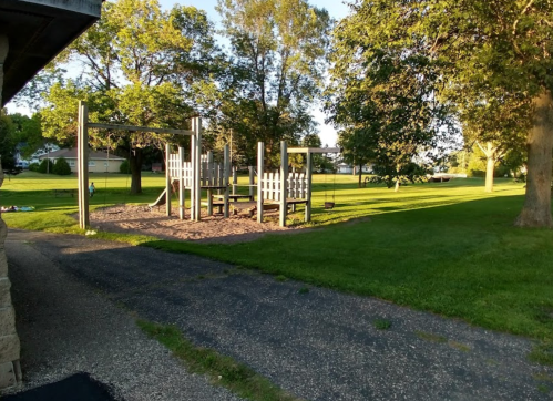 A playground with swings and climbing structures, surrounded by green grass and trees in a sunny park.