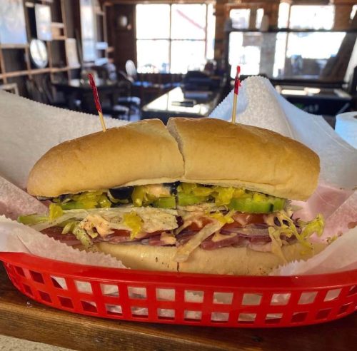 A large sandwich with layers of meats, vegetables, and sauces, served in a red basket with toothpicks on top.