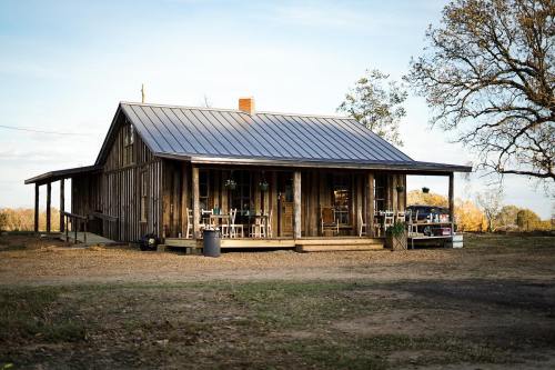 A rustic wooden house with a metal roof, surrounded by trees and a gravel path, featuring a porch and outdoor seating.
