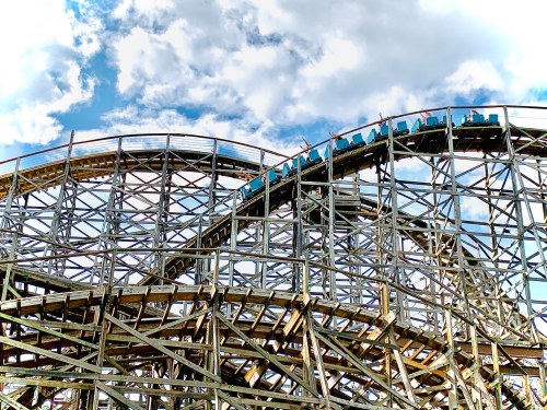 A wooden roller coaster with riders ascending a steep hill against a backdrop of blue sky and fluffy clouds.