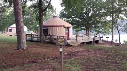 A wooden sign labeled "Yurt Two" in front of a yurt on a deck by a lake, surrounded by trees.