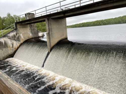 A dam with water flowing over its edge into a calm lake, surrounded by greenery under a cloudy sky.