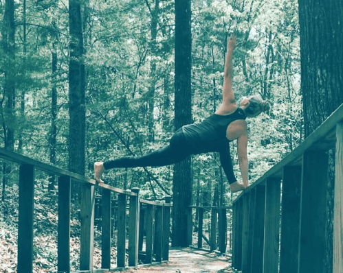 A person performs a yoga pose on a wooden railing in a forested area, surrounded by trees.