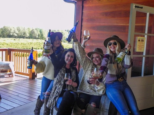 Four women celebrate outdoors, holding wine bottles and glasses, smiling and enjoying a sunny day on a wooden deck.