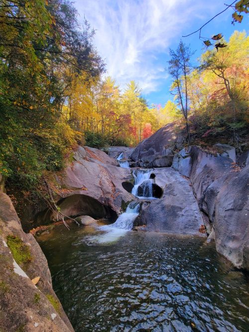 A serene waterfall cascades over rocks, surrounded by vibrant autumn foliage and a clear blue sky.