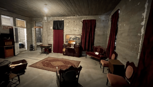 A dimly lit room with concrete walls, red curtains, vintage furniture, and a decorative rug.